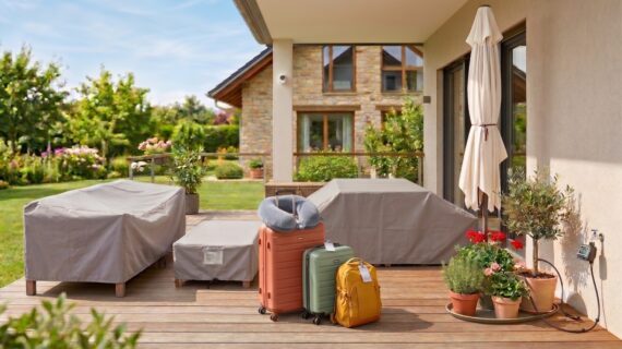 Eine sonnige Terrasse mit abgedeckten Gartenmöbeln und gepackten Koffern davor.